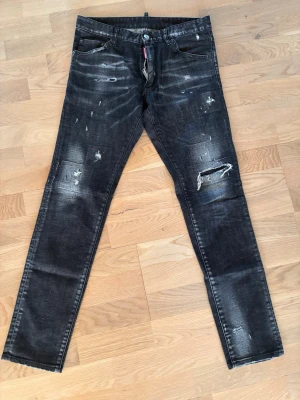 Svarta distressed jeans från Dsquared2 - Säljer ett par svarta jeans från Dsquared2 med slitna detaljer, hål och färgstänk. Jeansen har en smal passform, klassisk femficksdesign och dragkedja med Dsquared2-logga. Perfekta för dig som gillar en edgy och trendig stil.