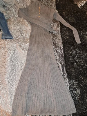 Grå ribbstickad långklänning - Snygg grå långklänning i ribbstickat material med kabelstickat mönster upptill. Klänningen har lång ärm, hög krage och dekorativa guldknappar framtill. Materialet är mjukt och stretchigt, perfekt för en trendig och bekväm look.