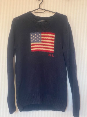 Mörkblå stickad tröja Polo Ralph Lauren - Mörkblå stickad tröja från Polo Ralph Lauren med amerikansk flagga i rött, vitt och blått på bröstet samt RL-broderi. Tröjan har rund halsringning, långa ärmar och ribbstickade muddar. Klassisk och tidlös design i mjukt material.