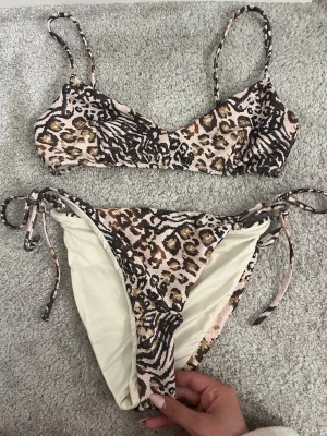 Leopardmönstrad bikini med knyt - Snygg bikini med leopardmönster i beige, brunt och svart. Överdelen har smala axelband och underdelen har knyt i sidorna för justerbar passform. Perfekt för strandhäng eller poolparty.