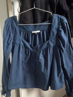 Blå långärmad blus från American Eagle - Snygg blå blus från American Eagle med lång ärm och smockade muddar. Blusen har knappar framtill, en djup fyrkantig ringning. Köpt i NYC för 500kr. Endast använd 2 gånger. 