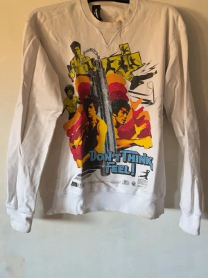 Vit sweatshirt med färgglatt tryck - Cool vit sweatshirt med stort grafiskt tryck på framsidan i gult, rött, svart och blått. Texten 'DON'T THINK FEEL!' står i blått framtill. Tröjan har rund hals och långa ärmar, perfekt för en avslappnad streetstyle-look.