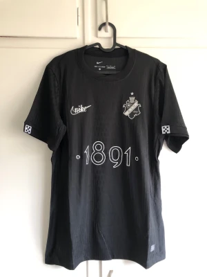 Svart AIK tröja från Nike - Snygg svart AIK t-shirt från Nike med klubbmärke på bröstet, vit text '1891' framtill och diskreta rutdetaljer på ärmarna. Tillverkad i lätt och bekvämt material, perfekt för träning eller matchdag.