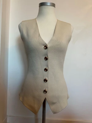 Beige ribbad väst med knappar - Stilren beige väst i ribbat material med djup v-ringning och sex stora dekorativa knappar framtill. Västen har en figurnära passform och spetsig nederkant som ger en modern look. Perfekt att styla över en skjorta eller t-shirt.