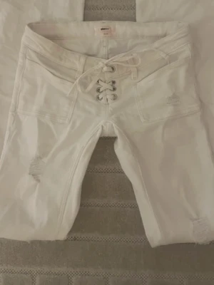 Vita snörade jeans med slitningar - Säljer ett par vita jeans med snörning i midjan och coola slitningar på benen. Jeansen har raka ben och stora fickor framtill. Perfekta för dig som vill ha en unik och trendig look.
