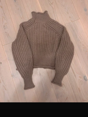 Brun ribbstickad polotröja - Säljer en brun ribbstickad polotröja med hög krage och ballongärmar. Tröjan har en croppad passform och är gjord i ett tjockare garn som känns mjukt och varmt. Perfekt för kyliga dagar och enkel att styla till jeans eller kjol.