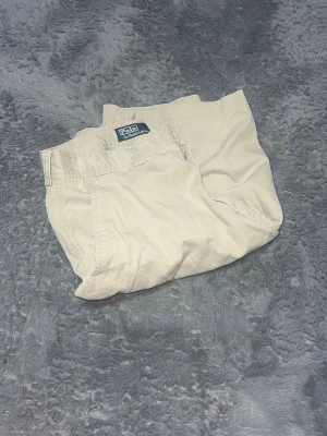 Polo Ralph Lauren Chino Cargo Shorts - Klassiska och praktiska cargoshorts i herrmodell från Polo Ralph Lauren i en tidlös beige färg. Shortsen är gjorda i en robust bomullskvalitet (chino-tyg) och har de karaktäristiska sidofickorna med lock. De har en avslappnad passform och är perfekta för sommarens alla äventyr. En rejäl och snygg baskomponent i garderoben. • Skick: Begagnat skick. Det finns en fläck på ena benet (se närbild på bild för en tydlig bild av fläcken). 