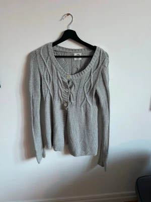 Grå kabelstickad kofta med knappar - Snygg grå kabelstickad kofta från H&M med tre stora knappar framtill. Koftan har rund halsringning, långa ärmar och ett klassiskt flätmönster upptill. Perfekt att slänga över en t-shirt för en chill look.