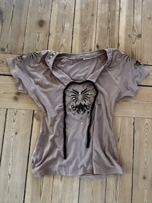 Beige topp med broderi från Angie - Säljer en beige topp från Angie med korta ärmar och dekorativt broderi framtill. Toppen har pärldetaljer vid halsringningen och ett unikt mönster i svart på bröstet. Perfekt för dig som gillar personliga detaljer och en avslappnad stil.