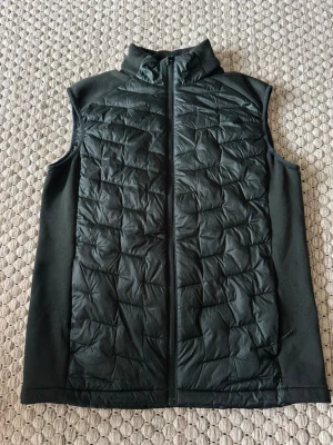 Mörkgrönt quiltad dunväst med dragkedja - Stilren mörkgrön dunväst med quiltat mönster och hög krage. Västen har hel dragkedja framtill och släta sidopaneler i stretchigt material för extra komfort. Perfekt lager på lager-plagg för kyliga dagar.