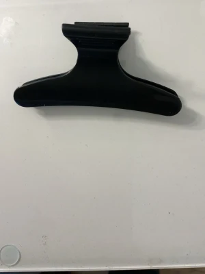 Svart hårklämma i plast - En enkel och stilren svart hårklämma i plast med klassisk fjärilsform. Perfekt för att snabbt sätta upp håret och hålla det på plats. Klämman har en matt finish och är lätt att använda för olika frisyrer.