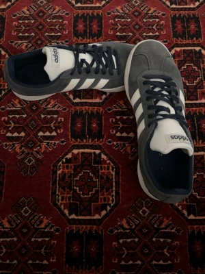 Grå Adidas sneakers med vita ränder - Snygga sneakers från Adidas i grå mocka med klassiska vita ränder på sidorna och vit sula. Skorna har mörkblå snören och detaljer samt Adidas-logga på plösen. Perfekta för en avslappnad streetstyle-look.
