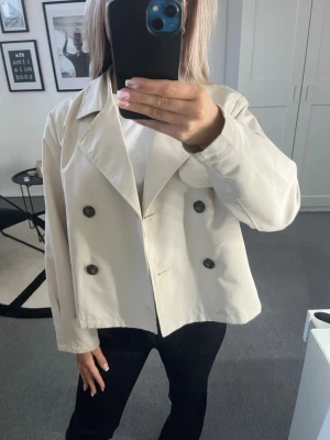 Beige kappa  - Snygg  kappa/trenchcoat i beige med klassisk dubbelknäppning och bred krage. Kappan har en rak passform och stora knappar framtill. Perfekt för dig som vill en klassisk look. Materialet är ganska tunt då den är bra till våren och sommaren. Den har lite smink fläckar på sig(se bild 4) men absolut inget som märks av!