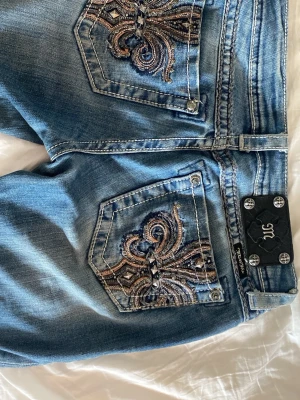 Flared Miss Me jeans  - Säljer ett par blå jeans från Miss Me med snygga broderade bakfickor och dekorativa detaljer. Jeansen har en klassisk femficksmodell, låg midja och utsvängda ben. Materialet är denim i bomull med slitningar för en cool look. Modellen är ”boot” men jag har sytt in kilar på båda sidor som gör byxorna mer flared (se sista bilden). Storlek 27, vilket motsvarar ungefär S. 