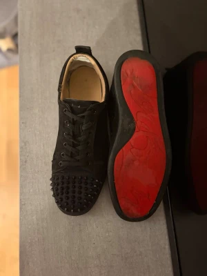 Svarta Louboutin sneakers med nitar - Säljer ett par svarta sneakers från Christian Louboutin med mattsvarta nitar på tån och den ikoniska röda sulan. Skorna har snörning och är tillverkade i mocka för en lyxig känsla. Perfekta för dig som vill sticka ut med en edgy och exklusiv stil. Jag har suttit dit lappat i hälen för att undervika slitage och tillfälligt sytt ihop lite på kanten för att få skon som ny igen där av det låga priset. Låda + kort följes med!