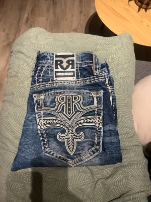 Rock Revival blå jeans med brodyr - Säljer ett par blå jeans från Rock Revival med grymt snygg vit brodyr på bakfickorna och tydliga kontrastsömmar. Jeansen har en klassisk femficksmodell och är gjorda i bomull med lite stretch för extra komfort. Perfekta för dig som vill sticka ut med detaljer!