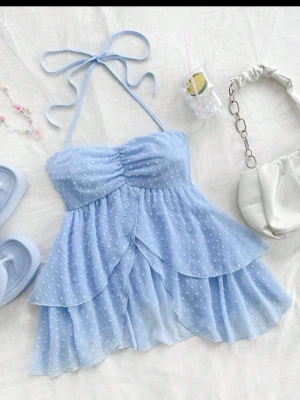 Ljusblå prickig volangtop - Supersöt ljusblå topp med volanger och prickigt mönster. Axelbandslös modell med knyt i nacken och smock baktill för skön passform. Tunn, luftig och flowy i syntetmaterial, perfekt för varma dagar. Ger en romantisk och trendig vibe.