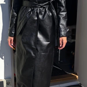Svart lång trenchcoat i skinnimitation.S.oanvänd - Helt ny oanvänd.Stilren svart trenchcoat i skinnimitation med bälte i midjan och klassisk krage. Kappa Långa ärmar med knappar vid ärmslut och slät yta ger en cool look. Perfekt för dig som vill ha en edgy och trendig jacka till hösten.storlek S. Kan även passa xs och M. Hämtas eller fraktas så fort så möjligt pågrund av flytt. Oanvänd helt ny.tror den är från shin