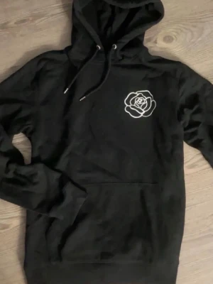 Svart hoodie med ros - Svart hoodie med stor huva och vitt tryck av en ros. Hoodien har magficka och ribbade muddar. Perfekt för dig som gillar streetwear och grafiska tryck. Aldrig använd, från trycklagret 🌹✨