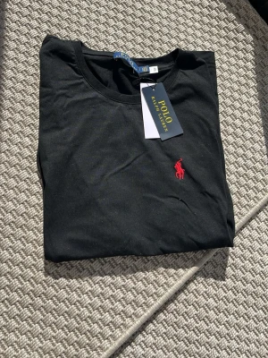 Svart t-shirt från Polo Ralph Lauren - Klassisk svart t-shirt från Polo Ralph Lauren med röd broderad logga på bröstet. T-shirten har rund halsringning och korta ärmar. Tillverkad i mjuk bomull som känns skön mot huden. Perfekt basplagg med stilren design.