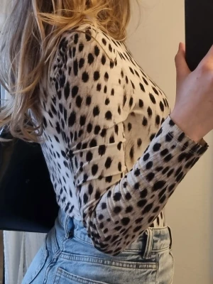 Djurmönstrad långärmad body i syntet - Säljer en slim, långärmad topp med svart prickigt ”djurmönster” på vit botten. Toppen har hög hals och är tillverkad i ett stretchigt syntetmaterial som sitter tight mot kroppen. Perfekt att styla med både jeans och skinnbyxor.