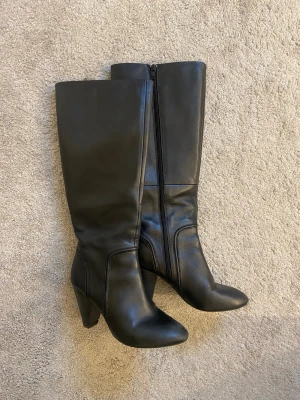 Svarta knähöga boots med klack - Säljer ett par svarta knähöga boots i slätt skinnimitation. Använda två gånger men de har inga defekter som påverkar helhetsupplevelsen! Kedjan funkar som den ska, inga repor eller annat. Har ingen aning vilken skobutik de är ifrån har tyvärr glömt🙈Pris går att diskutera😊
