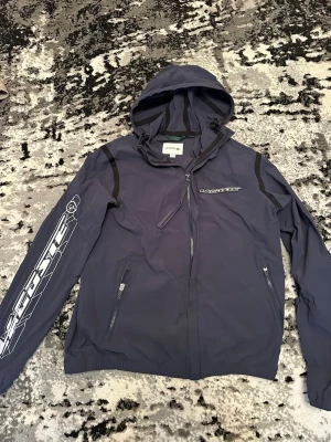Mörkblå vindjacka från Lacoste - Snygg mörkblå vindjacka från Lacoste med huva och dragkedja framtill. Jackan har två fickor med dragkedja, svarta detaljer på axlarna och stort Lacoste-tryck på bröstet och ärmen. Tillverkad i lätt och tåligt material, perfekt för blåsiga dagar.