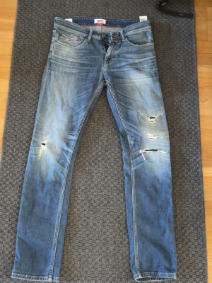Blå slitna jeans från Tommy Jeans - Ett par blå denimjeans från Tommy Jeans med slitningar och hål på framsidan. Jeansen har smal passform, klassisk femficksdesign och normal midja. Snygg tvättad look och tydliga slitdetaljer för en trendig stil.