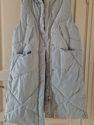 Ljusblå lång dunväst med huva - Lång ljusblå dunväst med quiltat mönster och huva. Västen har dragkedja och knappar framtill samt två stora fickor. Perfekt att slänga över en hoodie för extra värme. Materialet är syntet och dun, vilket gör den både lätt och varm.