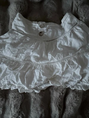 Vit volangkjol med knappar - Supersöt vit kjol med volanger och dekorativa sömmar. Kjolen har två knappar i midjan och resår baktill för skön passform. Tillverkad i ett lätt tyg, perfekt för sommardagar. Volangdetaljerna ger en romantisk och trendig look.