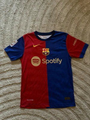 FC Barcelona matchtröja Nike - Snygg FC Barcelona matchtröja i rött och blått med guldfärgade detaljer. Tröjan har klubbmärke, Nike-logga och Spotify som sponsor framtill samt UNHCR-logga och nummer 19 med Lamine Yamal på ryggen. Tillverkad i lätt och ventilerande material.