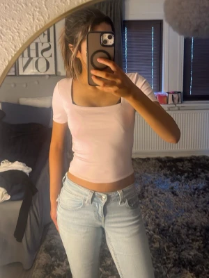 Ljusrosa croppad topp - En ljusrosa croppad topp med korta ärmar och fyrkantig ringning. Toppen har en slim passform och är tillverkad i ett mjukt, stretchigt material som sitter tajt mot kroppen. Perfekt att styla med jeans eller kjol för en trendig look.