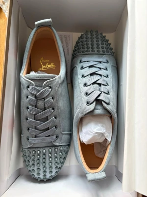 Gråa Christian Louboutin sneakers - Snygga grå sneakers från Christian Louboutin med ikoniska röda sulor och nitar på tån. Skorna har en låg profil, snörning och är tillverkade i tyg med läderdetaljer. Perfekta för dig som vill sticka ut med exklusiv design och edgy detaljer.