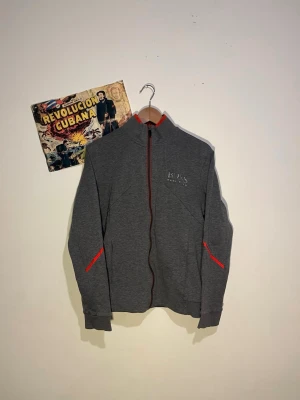 Grå dress från Hugo Boss - Snygg grå ziptröja från Hugo Boss med röd dragkedja och röda detaljer vid fickorna. Tröjan har hög krage och diskret Boss-logga broderad på bröstet. Perfekt för en avslappnad men stilren look. Snygga matchande byxor.