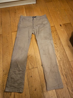 Beige chinos med rak passform - Snygga beige chinos med rak passform och klassisk design. Byxorna har knapp och dragkedja framtill samt bälteshällor. Perfekta för en stilren och avslappnad look. Materialet känns mjukt och lätt, vilket gör dem bekväma att bära hela dagen. I bilder ser man inte men baksidan har 2 fickor med svarta knappar till, måtten syns inte heller där breden är 42cm och längden är 105cm