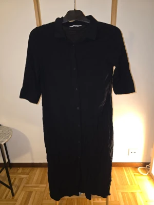 Svart skjortklänning från ONLY - Stilren svart skjortklänning från ONLY med knappar hela vägen framtill och klassisk krage. Klänningen har trekvartsärm och rak passform, perfekt för en enkel och clean look. Tillverkad i mjukt tyg som känns bekvämt mot huden.
