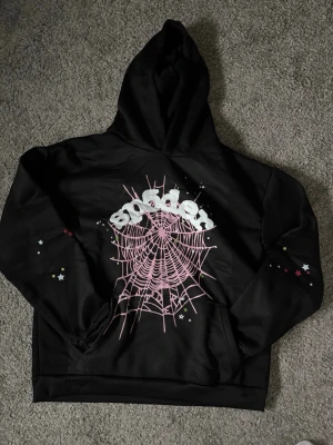 Svart hoodie med spindelnätsmotiv - Säljer en svart hoodie från Sp5der med stor rosa spindelnätsgrafik och vit text på ryggen. Hoodien har huva och ficka framtill samt små stjärndetaljer i flera färger på ärmar och rygg. Perfekt för dig som gillar streetwear och unika prints.