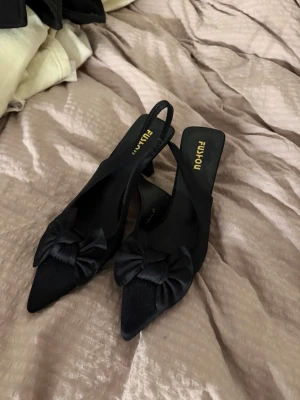 Svarta ballerinaskor med rosett - Stilrena svarta ballerinaskor från FUIYHOU med spetsig tå och dekorativa rosetter framtill. Skorna har öppen häl med rem och är tillverkade i ett tygmaterial som ger en elegant look. Perfekta för dig som gillar klassisk och trendig stil.