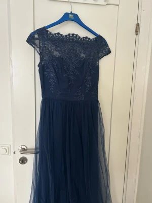 Marinblå långklänning med spets - Elegant marinblå långklänning med vacker spets upptill och transparent axelparti. Kjolen är i flera lager tunn mesh som ger ett svepande fall. Perfekt för speciella tillfällen där du vill känna dig extra fin.