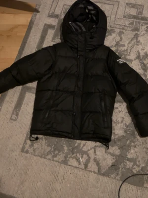 Svart pufferjacka  - Säljer en svart pufferjacka med huva och logga på ärmen. Jackan har dragkedja och knappar framtill samt justerbar nederkant med snören. Perfekt för kalla vinterdagar och riktigt skön att bära.