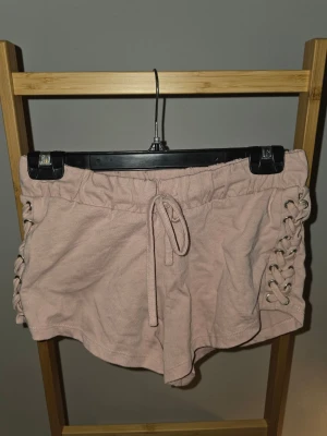Ljusrosa shorts med snörning i sidorna - Säljer ett par ljusrosa shorts med snörning i sidorna och resår i midjan. De har en avslappnad passform och är tillverkade i mjuk bomull, perfekta för varma dagar. Snygga detaljer som ger en sportig och trendig vibe.
