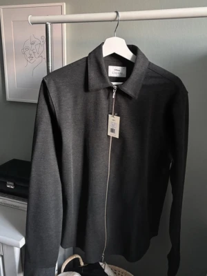 Mörkgrå overshirt från Ciszere - Nypris 1500kr. Snygg mörkgrå overshirt från Ciszere med rak passform och klassisk krage. Jackan har dragkedja framtill och är tillverkad i ett mjukt, slätt material som ger en clean look. Perfekt att bära över en t-shirt eller skjorta för en stilren vibe.