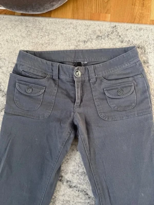 Mörkgrå byxor med fickdetaljer - Snygga mörkgrå byxor med två framfickor med lock och knappdetaljer. Byxorna har låg midja och är bootcut. Storlek 36, köpta second hand. Har små vita fläckar men de syns knappt!