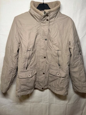 Beige vindjacka med hög krage - Stilren beige vindjacka med hög ribbad krage och dragkedja framtill. Jackan har flera fickor med både dragkedja och knappar, samt diskreta sömdetaljer. Perfekt för kyliga dagar och enkel att matcha med olika outfits.