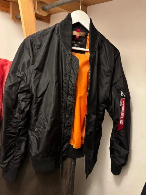 Svart bomberjacka från Alpha Industries - Säljer en klassisk svart bomberjacka från Alpha Industries med orange innerfoder och ikonisk röd 'Remove Before Flight'-tag på ärmen. Jackan har ribbade muddar, dragkedja framtill och fickor på sidorna samt ärmen. Perfekt för en streetwear-look. Använd en gång under en sommar, lite nopprig (se bild) men annars fin.