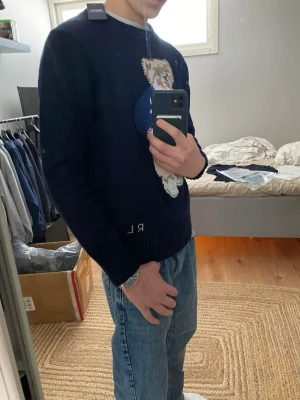 Mörkblå stickad tröja med björnmotiv - Säljer en mörkblå stickad tröja från Ralph Lauren med ett stort broderat björnmotiv på framsidan. Tröjan har rund halsringning, ribbstickade muddar och RL-broderi nedtill. Perfekt för dig som gillar klassiska och lekfulla detaljer.