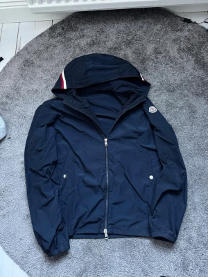 Mörkblå Moncler vindjacka med huva - Snygg mörkblå vindjacka från Moncler med huva och dragkedja framtill. Jackan har vita och röda detaljer på huvan samt Moncler-logga på ärmen. Två fickor med tryckknappar, perfekt nu när de börjar bli varmare.