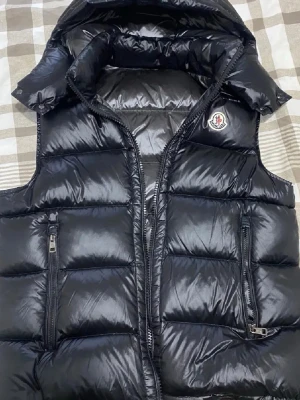 Svart dunväst från Moncler - Snygg svart dunväst från Moncler med glansig finish och två dragkedjeförsedda fickor framtill. Västen har hög krage och ikonisk Moncler-logga på bröstet. Perfekt för lager-på-lager och streetwear-vibe.