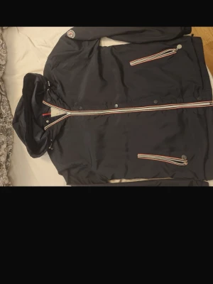 Marinblå vindjacka Moncler - Snygg marinblå vindjacka Moncler med huva och dragkedjor med röd, vit och blå detalj. Jackan har två fickor framtill och tryckknappar längs dragkedjan. Perfekt för dig som vill ha en stilren och sportig look.