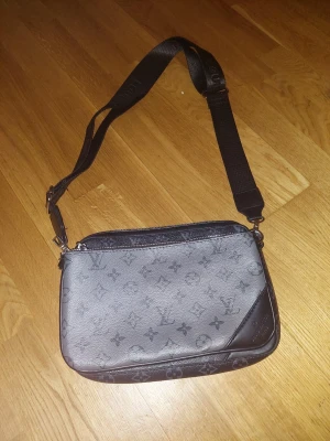 Louis Vuitton axelväska monogram svart - Snygg axelväska från Louis Vuitton i svart och grått med klassiskt monogrammönster. Väskan har justerbar axelrem i textil med Louis Vuitton-logga och detaljer i läder. Perfekt storlek för det viktigaste och stilren design som sticker ut.